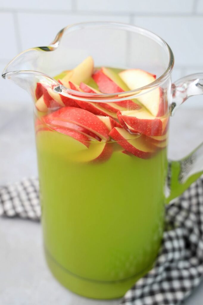 Grinch Christmas Punch with red apples in a glass pitcher on a gray plaid napkin.