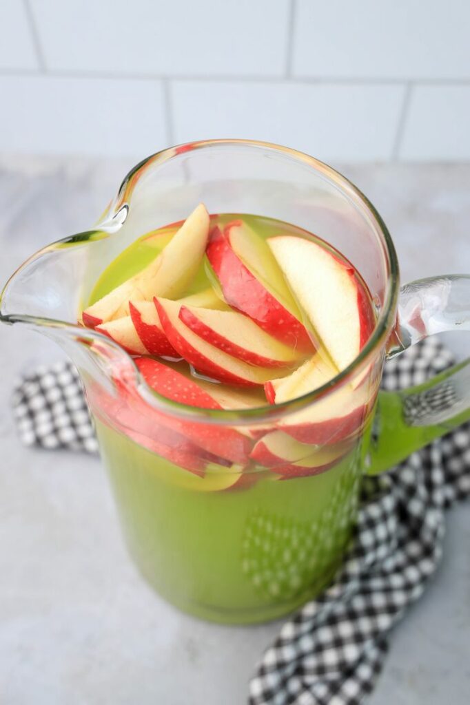 Grinch Christmas Punch with red apples in a glass pitcher on a gray plaid napkin.