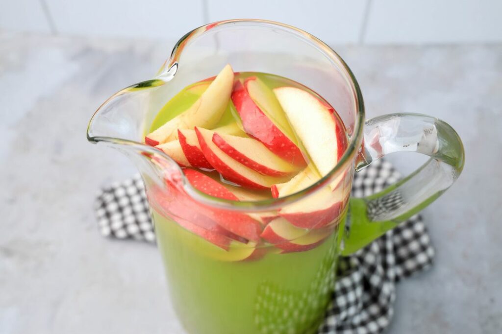 Grinch Christmas Punch with red apples in a glass pitcher on a gray plaid napkin.