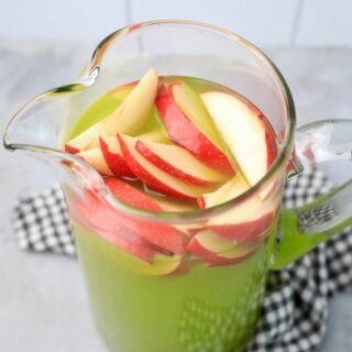 Grinch Christmas Punch with red apples in a glass pitcher on a gray plaid napkin.