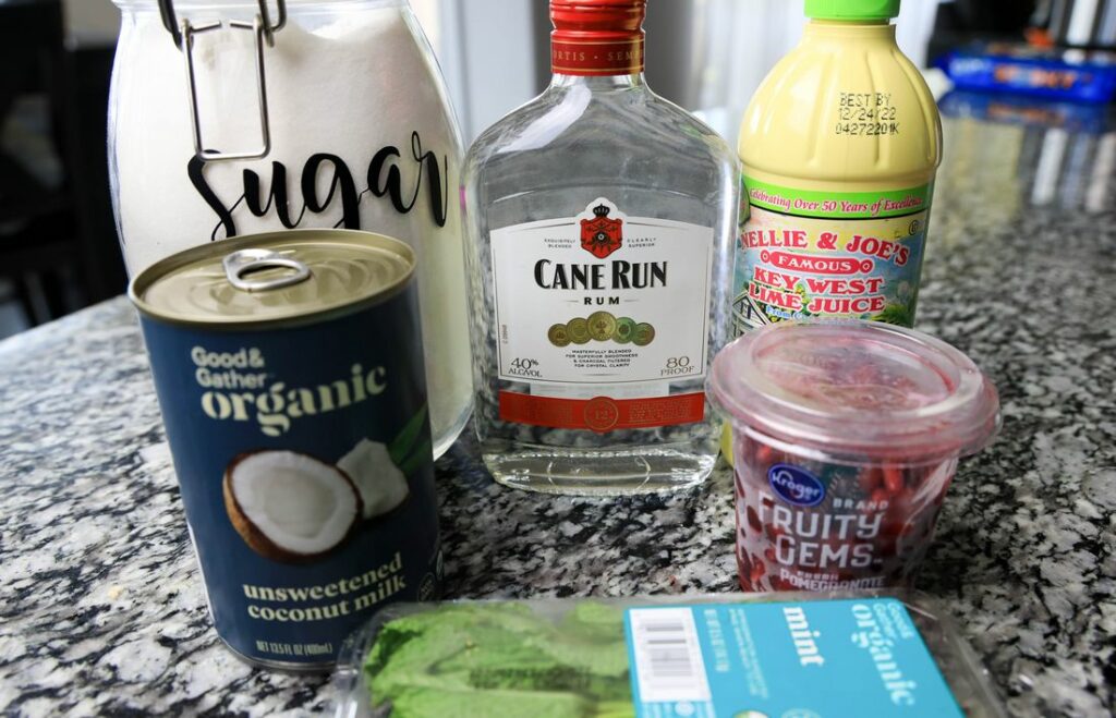 Rum, coconut milk, mint leaves, pomegranate, key lime juice, and sugar on a counter top.