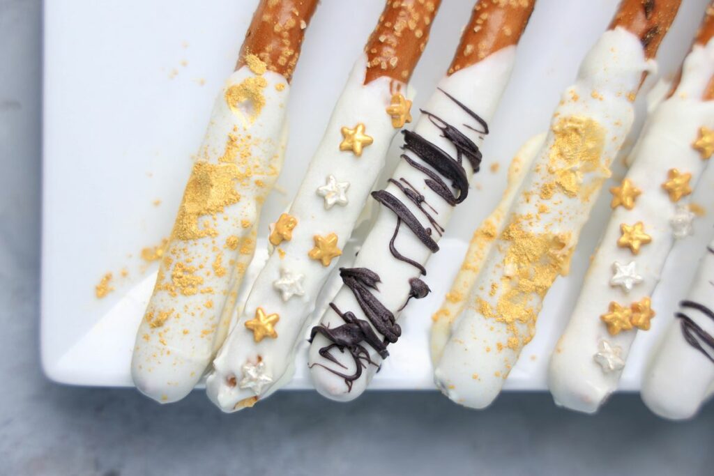 New Year's Eve Pretzel Rods dipped in chocolate with black drizzle and gold sprinkles on a white plate on a faux concrete backdrop.