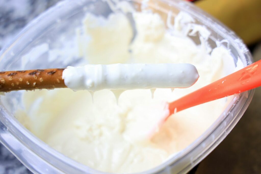 Pretzel rod with white chocolate hanging over a plastic bowl of white chocolate.