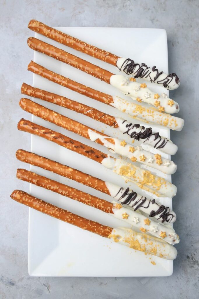 New Year's Eve Pretzel Rods dipped in chocolate with black drizzle and gold sprinkles on a white plate on a faux concrete backdrop.