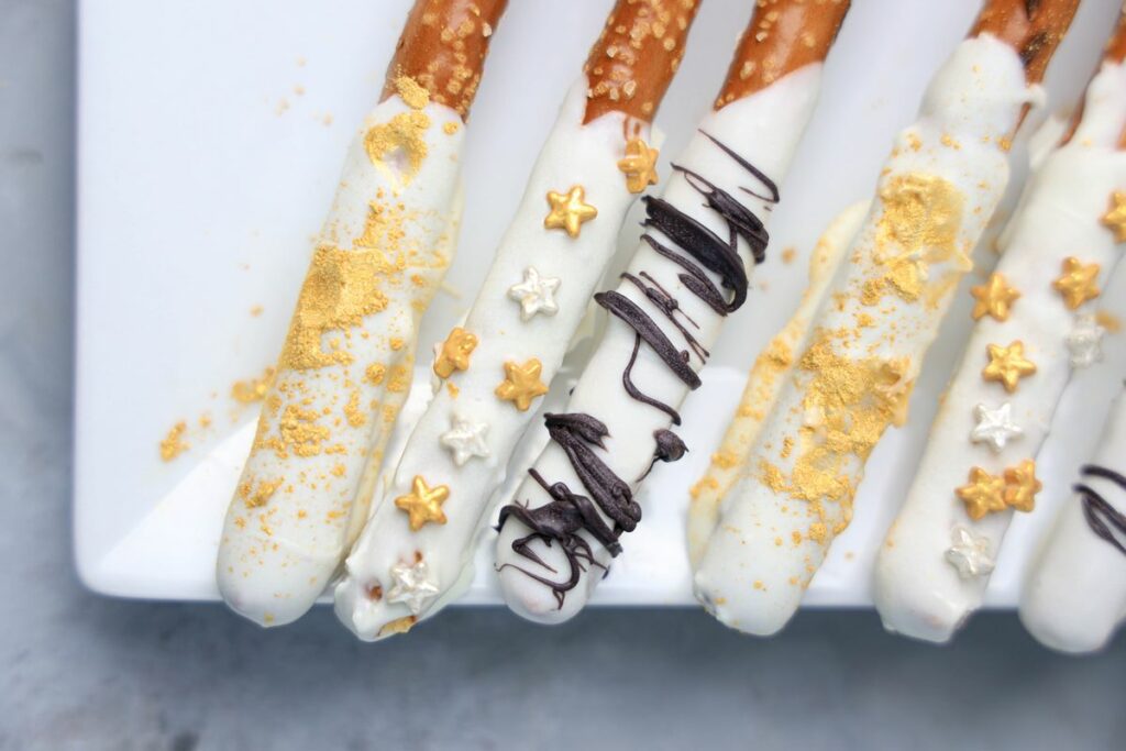 New Year's Eve Pretzel Rods dipped in chocolate with black drizzle and gold sprinkles on a white plate on a faux concrete backdrop.