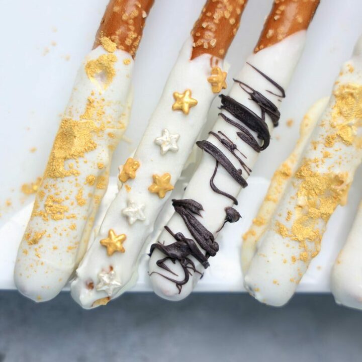 New Year's Eve Pretzel Rods dipped in chocolate with black drizzle and gold sprinkles on a white plate on a faux concrete backdrop.