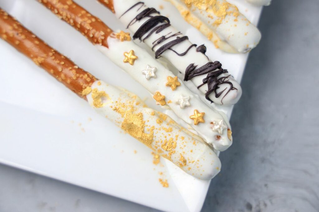 New Year's Eve Pretzel Rods dipped in chocolate with black drizzle and gold sprinkles on a white plate on a faux concrete backdrop.