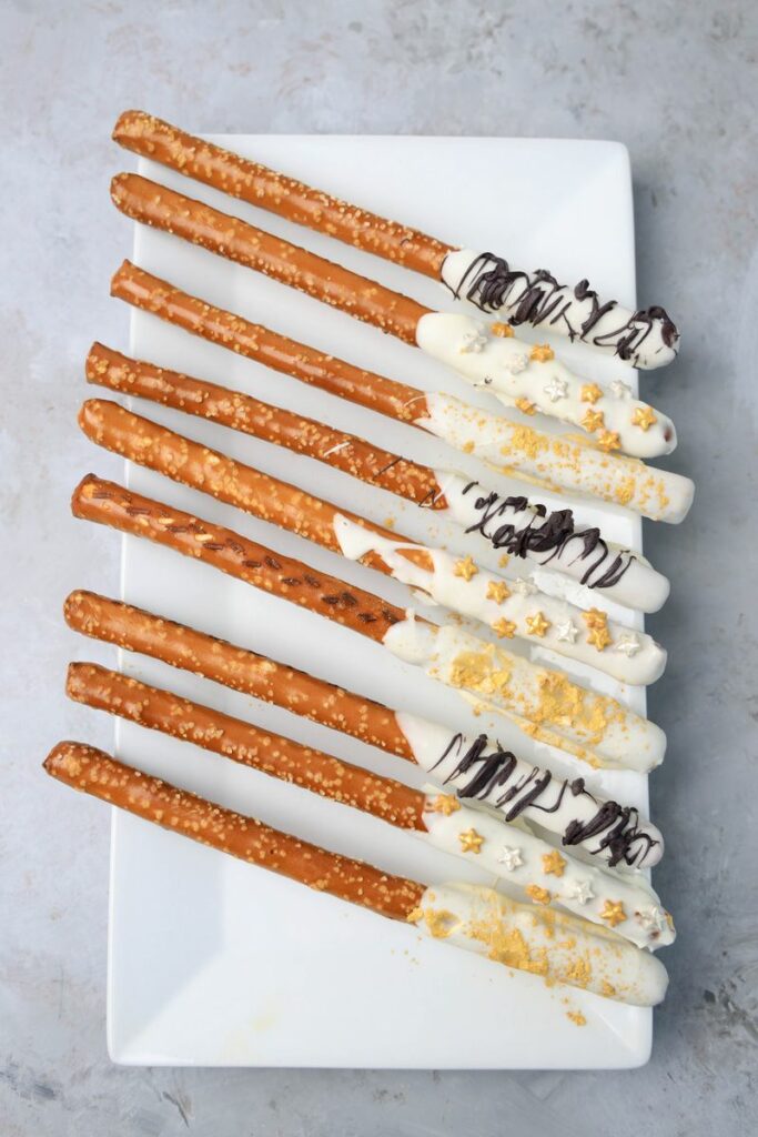 New Year's Eve Pretzel Rods dipped in chocolate with black drizzle and gold sprinkles on a white plate on a faux concrete backdrop.