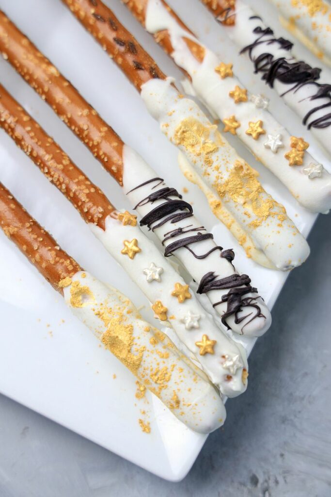 New Year's Eve Pretzel Rods dipped in chocolate with black drizzle and gold sprinkles on a white plate on a faux concrete backdrop.