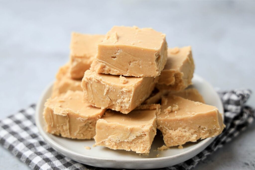 Peanut Butter Fudge stacked on a white plate on a gray plaid napkin with a faux concrete backdrop.