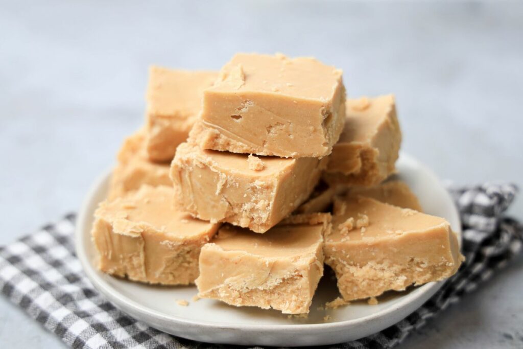 Peanut Butter Fudge stacked on a white plate on a gray plaid napkin with a faux concrete backdrop.