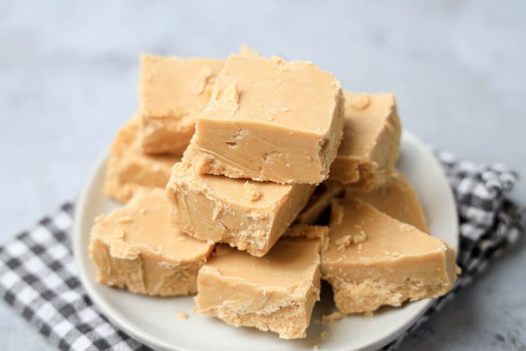 Peanut Butter Fudge stacked on a white plate on a gray plaid napkin with a faux concrete backdrop.