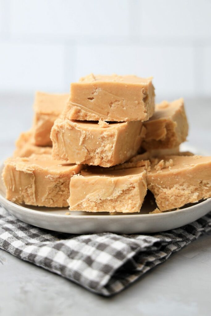 Peanut Butter Fudge stacked on a white plate on a gray plaid napkin with a faux concrete backdrop.