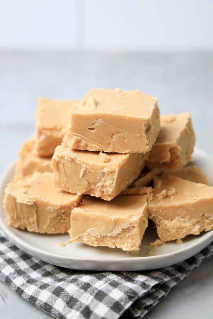 Peanut Butter Fudge stacked on a white plate on a gray plaid napkin with a faux concrete backdrop.