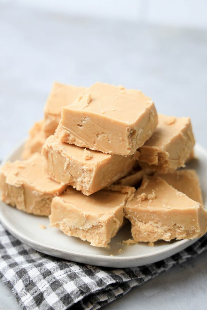 Peanut Butter Fudge stacked on a white plate on a gray plaid napkin with a faux concrete backdrop.