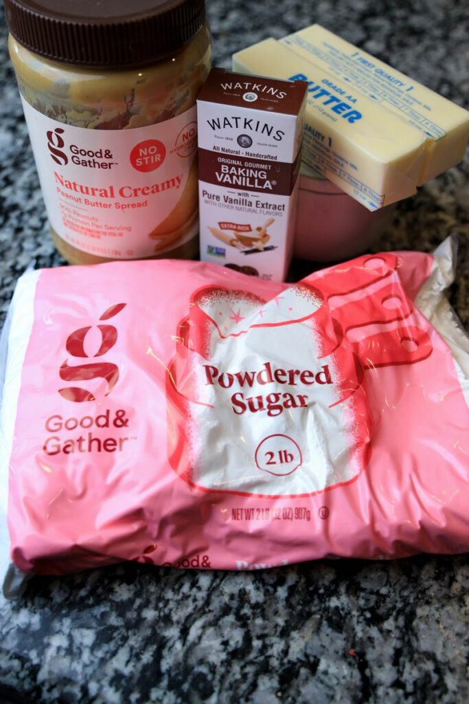 Ingredients to make peanut butter fudge. Powdered sugar, peanut butter, vanilla, and butter on a marble counter top.