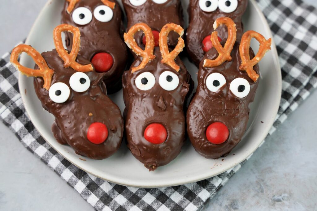 Rudolph Nutter Butters dipped in dark chocolate with a red M&M nose, candy eyes, and pretzel antlers on a white plate with a gray plaid napkin and a faux concrete backdrop.