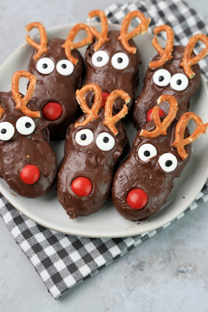 Rudolph Nutter Butters dipped in dark chocolate with a red M&M nose, candy eyes, and pretzel antlers on a white plate with a gray plaid napkin and a faux concrete backdrop.