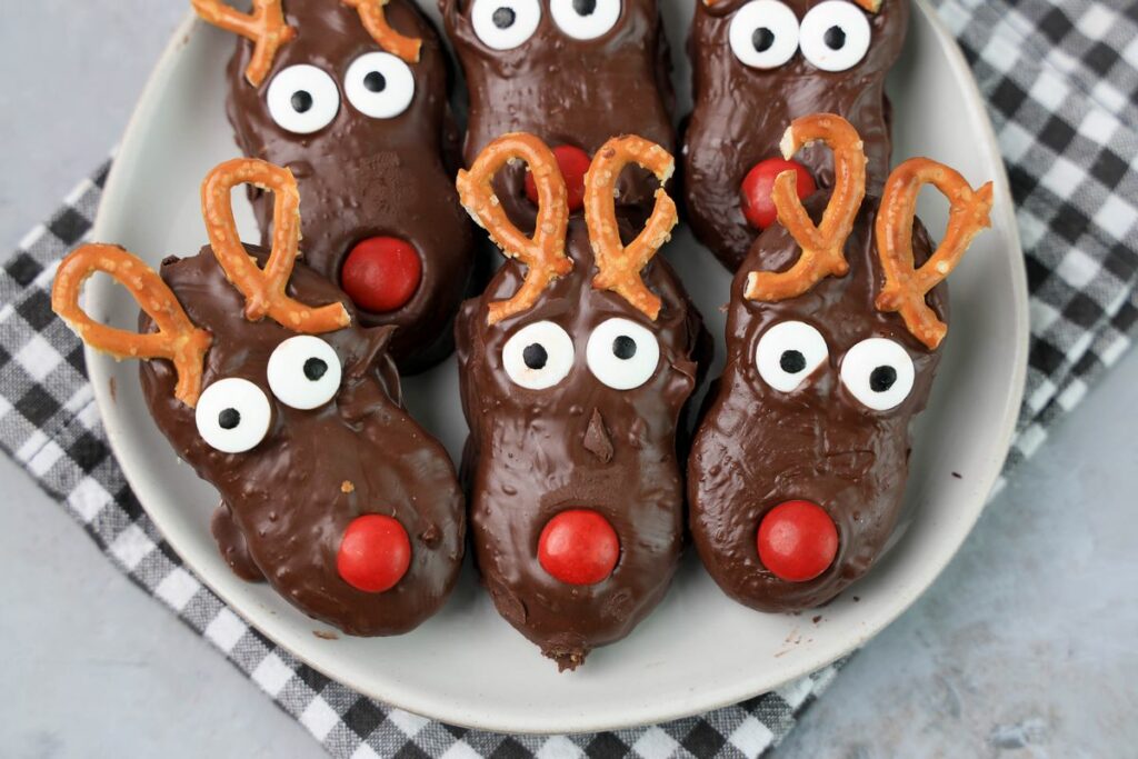 Rudolph Nutter Butters dipped in dark chocolate with a red M&M nose, candy eyes, and pretzel antlers on a white plate with a gray plaid napkin and a faux concrete backdrop.