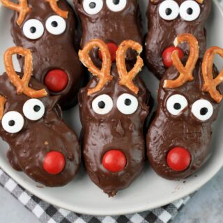 Rudolph Nutter Butters dipped in dark chocolate with a red M&M nose, candy eyes, and pretzel antlers on a white plate with a gray plaid napkin and a faux concrete backdrop.
