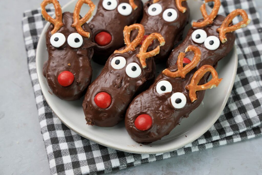 Rudolph Nutter Butters dipped in dark chocolate with a red M&M nose, candy eyes, and pretzel antlers on a white plate with a gray plaid napkin and a faux concrete backdrop.