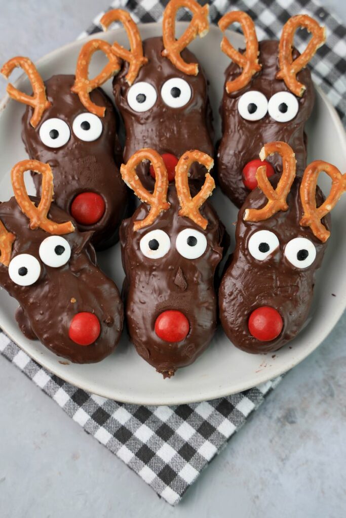 Rudolph Nutter Butters dipped in dark chocolate with a red M&M nose, candy eyes, and pretzel antlers on a white plate with a gray plaid napkin and a faux concrete backdrop.