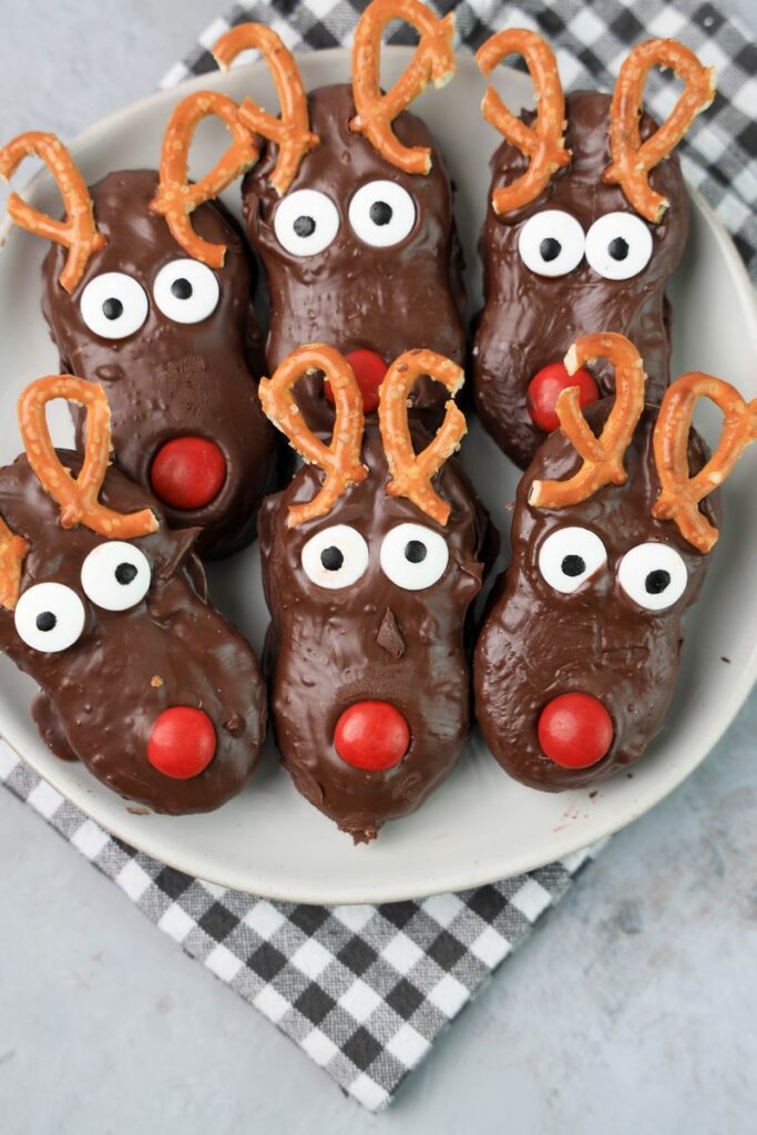 Rudolph Nutter Butters dipped in dark chocolate with a red M&M nose, candy eyes, and pretzel antlers on a white plate with a gray plaid napkin and a faux concrete backdrop.