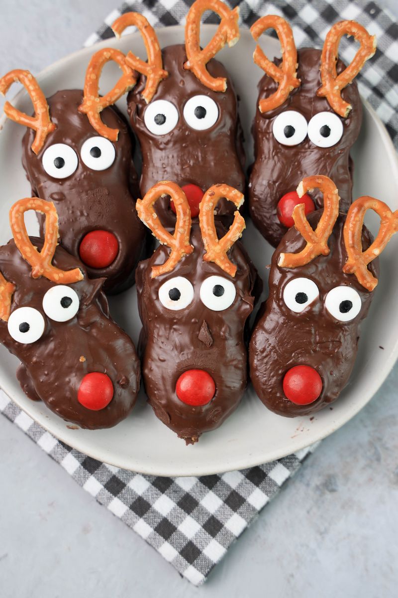 Rudolph Nutter Butters dipped in dark chocolate with a red M&M nose, candy eyes, and pretzel antlers on a white plate with a gray plaid napkin and a faux concrete backdrop.
