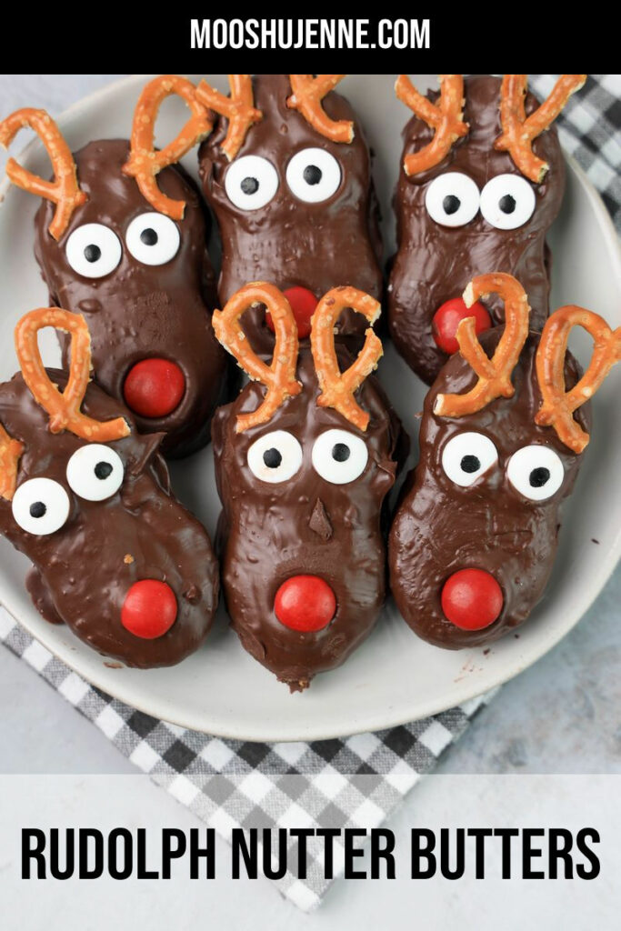 Rudolph Nutter Butters dipped in dark chocolate with a red M&M nose, candy eyes, and pretzel antlers on a white plate with a gray plaid napkin and a faux concrete backdrop.