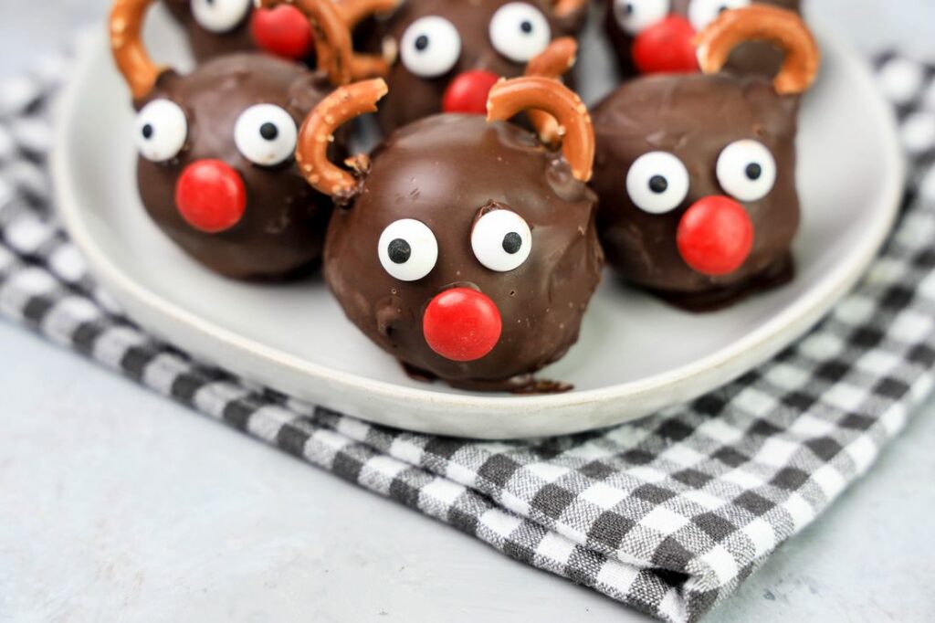 Rudolph orep balls with candy eyes, a red nose, and pretzel antlers on a white plaid with gray plaid napkin and faux concrete backdrop.