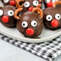 Rudolph orep balls with candy eyes, a red nose, and pretzel antlers on a white plaid with gray plaid napkin and faux concrete backdrop.