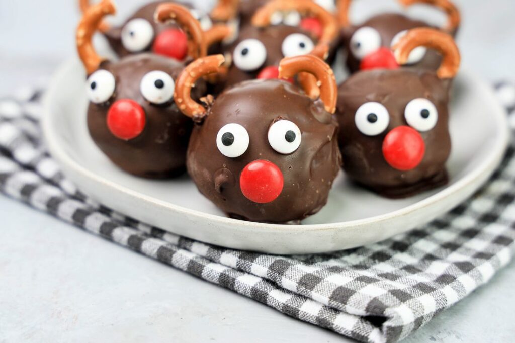 Rudolph orep balls with candy eyes, a red nose, and pretzel antlers on a white plaid with gray plaid napkin and faux concrete backdrop.