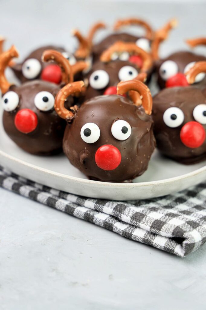 Rudolph orep balls with candy eyes, a red nose, and pretzel antlers on a white plaid with gray plaid napkin and faux concrete backdrop.