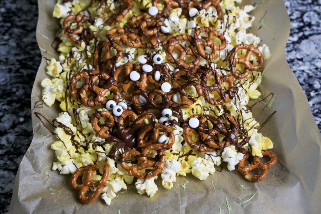 Popcorn and pretzels with chocolate and candy eyes on a baking sheet.