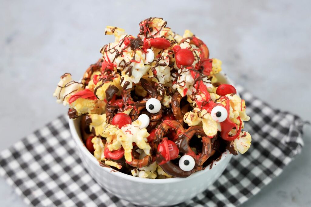 Popcorn with pretzels, red M&M's, chocolate and red drizzle in a white bowl.