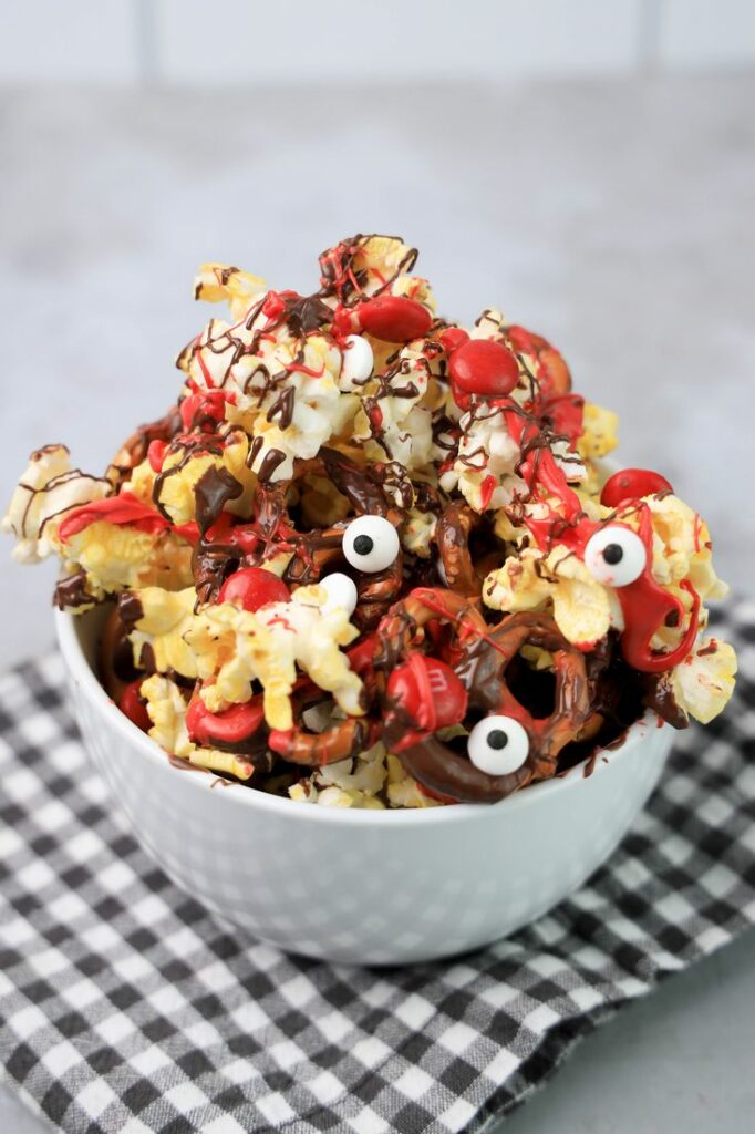 Popcorn with pretzels, red M&M's, chocolate and red drizzle in a white bowl.