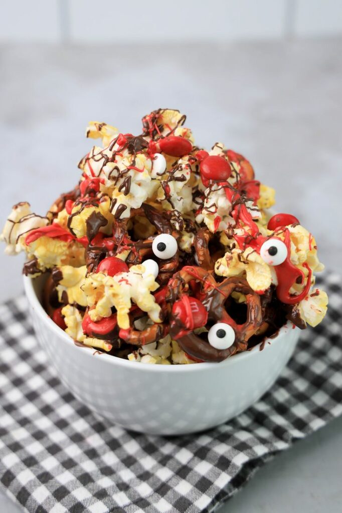 Popcorn with pretzels, red M&M's, chocolate and red drizzle in a white bowl.