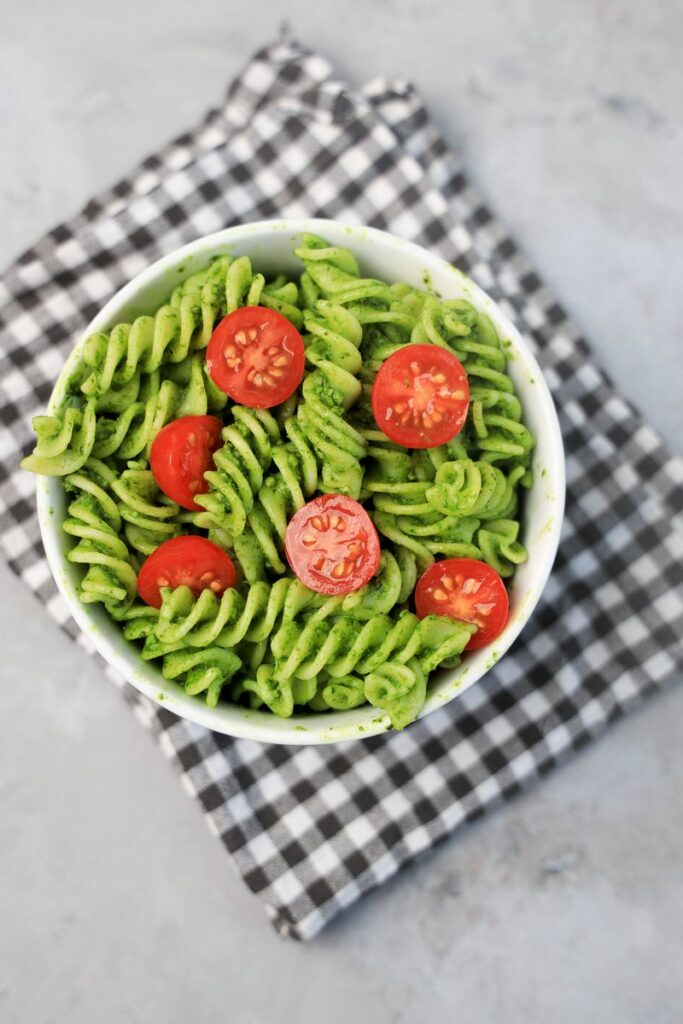 Basil Garlic Spinach Pesto Rotini topped with tomatoes in a white bowl on a gray plaid napkin.