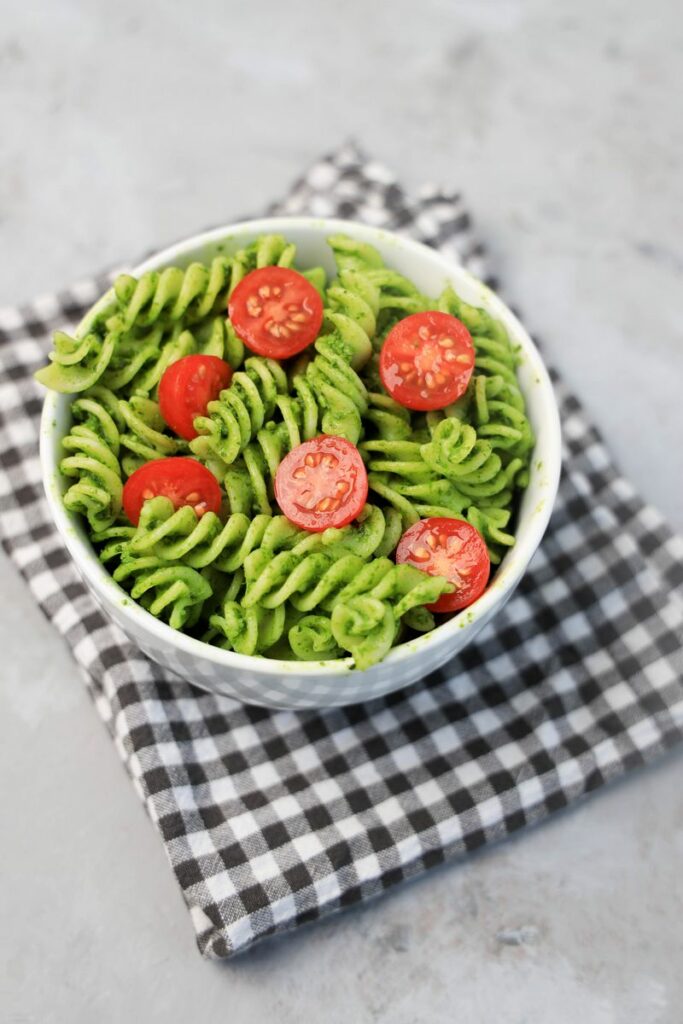 Basil Garlic Spinach Pesto Rotini topped with tomatoes in a white bowl on a gray plaid napkin.