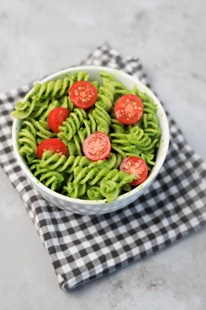 Basil Garlic Spinach Pesto Rotini topped with tomatoes in a white bowl on a gray plaid napkin.