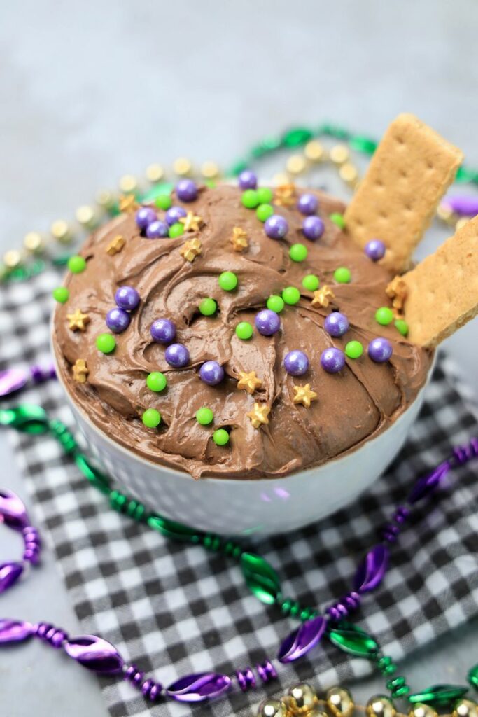 Mardi Gras Brownie Dip in a white bowl on a gray plaid napkin with mardi gras beads on a faux concrete backdrop.