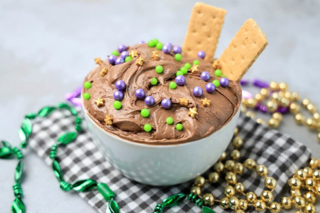 Mardi Gras Brownie Dip in a white bowl on a gray plaid napkin with mardi gras beads on a faux concrete backdrop.