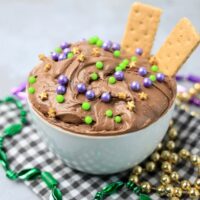 Mardi Gras Brownie Dip in a white bowl on a gray plaid napkin with mardi gras beads on a faux concrete backdrop.