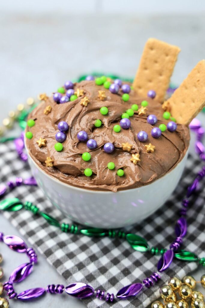 Mardi Gras Brownie Dip in a white bowl on a gray plaid napkin with mardi gras beads on a faux concrete backdrop.