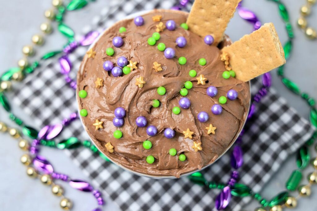 Mardi Gras Brownie Dip in a white bowl on a gray plaid napkin with mardi gras beads on a faux concrete backdrop.