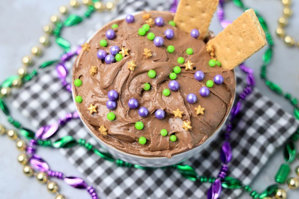 Mardi Gras Brownie Dip in a white bowl on a gray plaid napkin with mardi gras beads on a faux concrete backdrop.