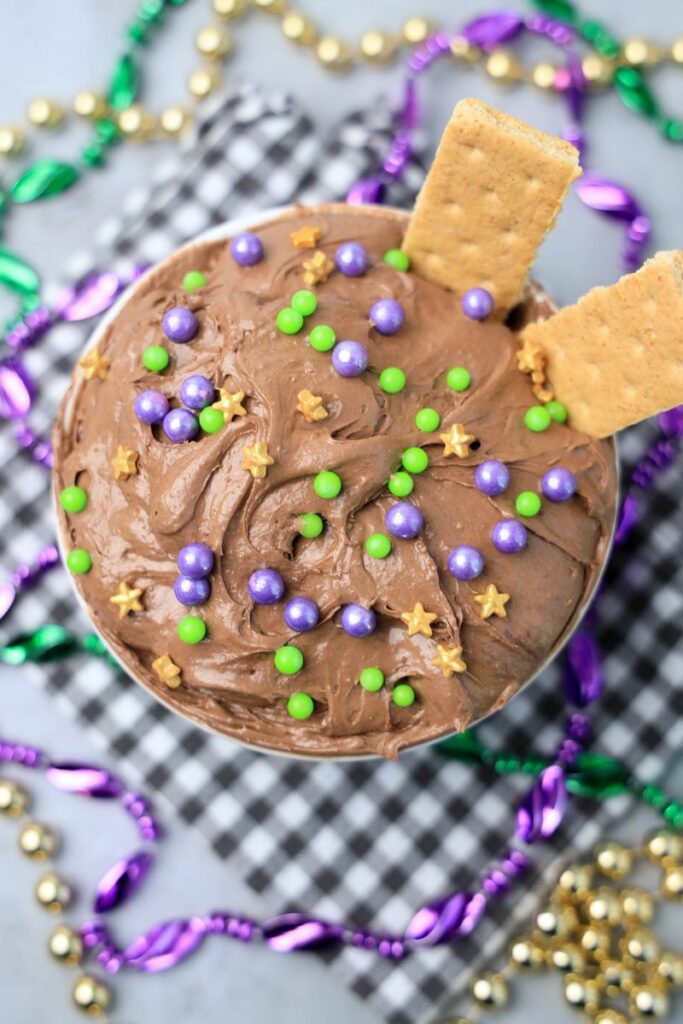 Mardi Gras Brownie Dip in a white bowl on a gray plaid napkin with mardi gras beads on a faux concrete backdrop.