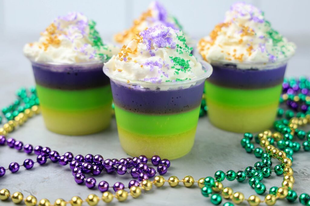 Mardi Gras Jello Shots layered with yellow, green, and purple. On a faux concrete backdrop with Mardi Gras Beads.