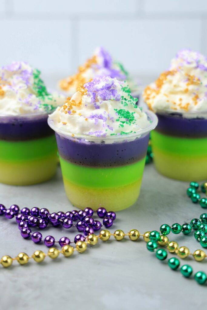 Mardi Gras Jello Shots layered with yellow, green, and purple. On a faux concrete backdrop with Mardi Gras Beads.