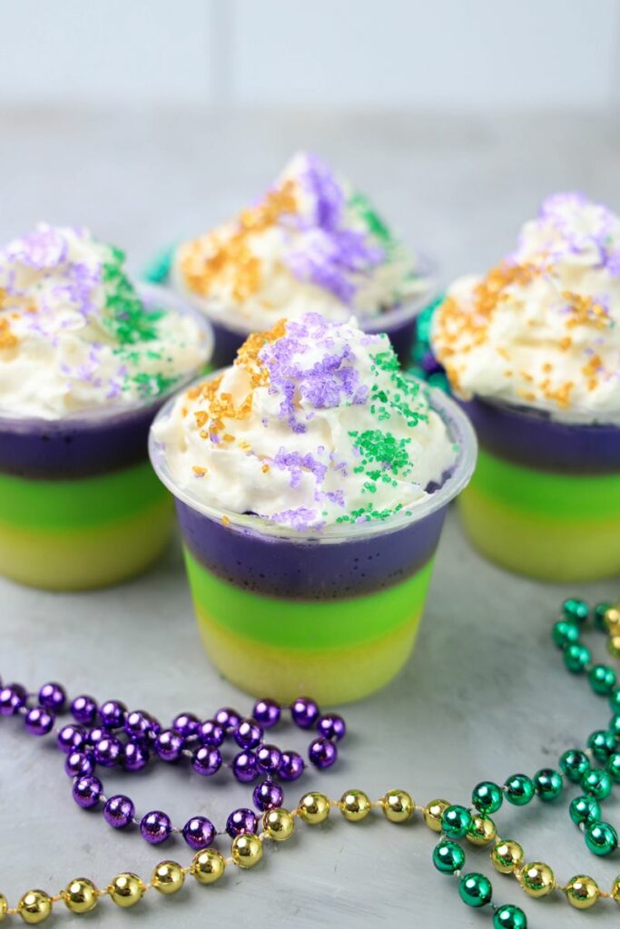 Mardi Gras Jello Shots layered with yellow, green, and purple. On a faux concrete backdrop with Mardi Gras Beads.